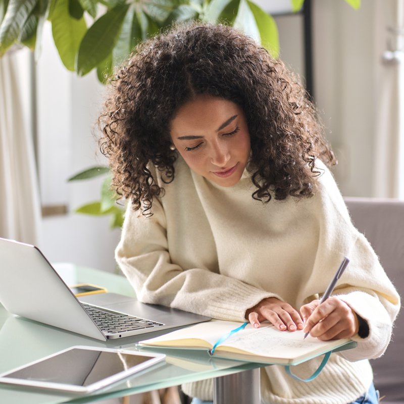 Woman writing in a notebook next to a laptop.